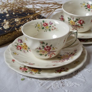 May include: Cream-colored teacups and saucers with a floral pattern featuring red, yellow, and purple roses. The teacups have a curved handle and sit on matching saucers and plates. The set is arranged on a white tablecloth, suggesting a vintage tea set.