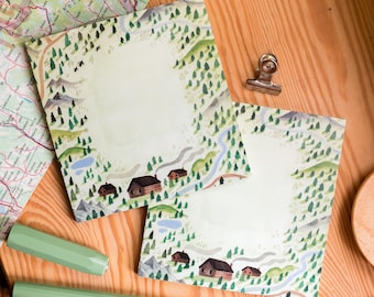 May include: Two green and white notepad sets with a woodland scene border. The border features trees, a river, and small cabins. The notepads are blank and ready to be written on.