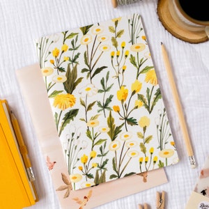 May include: A white notebook with a floral pattern featuring yellow and white flowers on a white background. The notebook is lying on a white surface with a yellow pencil and a wooden coaster with a coffee cup.