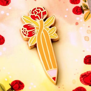 Könnte beinhalten: Ein goldfarbener Emaille-Pin in Form eines Bleistifts mit roten Blumen und gelben Blättern. Der Bleistift hat eine rosa Radiergummispitze und gelbe Streifen. Der Pin ist vor einem floralen Hintergrund mit roten Blumen und grünen Blättern platziert.