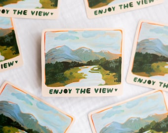 May include: Square stickers with a landscape painting of mountains, a river, and trees. The stickers have an orange border and the words "ENJOY THE VIEW" in black. The stickers are scattered on a white surface.