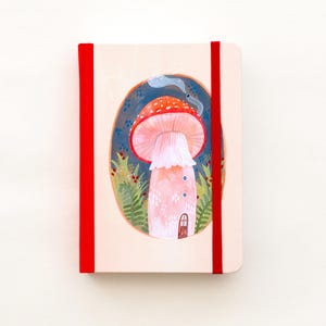 Può includere: Un taccuino con un'illustrazione di un fungo. La copertina presenta un fungo con cappello rosso e gambo rosa, una piccola porta e fumo. Un elastico rosso assicura il taccuino.