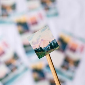 May include: A small, hand-painted watercolor painting of a mountain scene with a pink sky and green trees. The painting is mounted on a wooden stick.