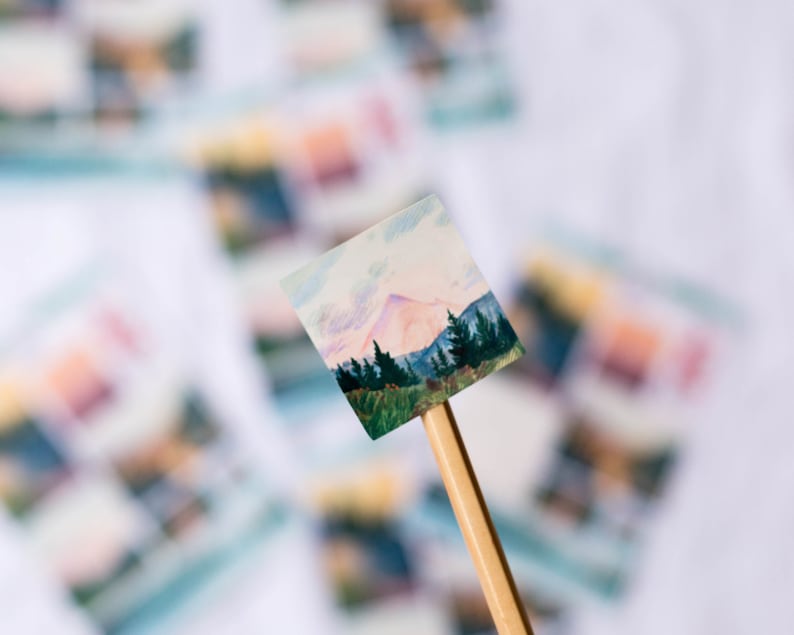 May include: A small, hand-painted watercolor painting of a mountain scene with a pink sky and green trees. The painting is mounted on a wooden stick.