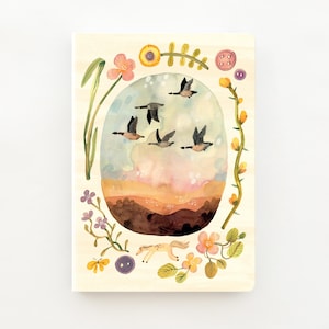 Könnte beinhalten: Ein rechteckiges Notizbuch mit einer Aquarellillustration von fünf Gänsen, die über eine Hügellandschaft und einen Sonnenuntergangshimmel fliegen. Die zentrale ovale Szene ist von bunten handgemalten Blumen, grünen Ranken und Knöpfen umrahmt.