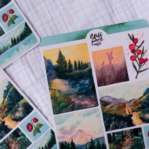 May include: A sheet of stickers with a watercolor aesthetic featuring nature scenes. The stickers include a deer in a field, a mountain range, a forest path, a river, and a berry branch. The stickers are labeled "Wild Pencil".