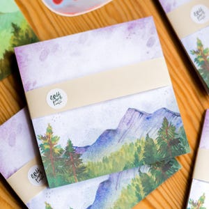 Può includere: Pile di blocchi per appunti quadrati con scene di montagne e foreste ad acquerello. Ogni blocco per appunti è avvolto con una fascia beige e un piccolo adesivo rotondo con la scritta "wild pencil". Lo sfondo presenta un lavaggio ad acquerello viola e bianco.