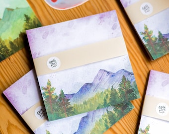 May include: Several square notepads featuring watercolour mountain and forest designs. Each pad is wrapped with a beige band and a small round sticker with the text "wild pencil". The background has a purple and white watercolour wash.