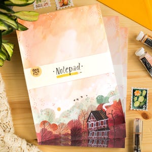 May include: A notepad with a watercolour landscape design featuring a house, trees, and birds. The notepad has the word "Notepad" printed on it. The colour palette includes shades of orange, pink, and brown.