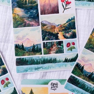 May include: A set of stickers featuring watercolor illustrations of forest scenes, including a mountain range, a lake, and a path through the woods. The stickers are printed on white paper and have a glossy finish.