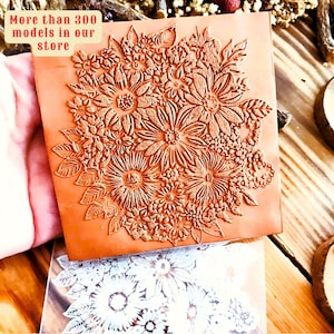 May include: A close-up of a brown rubber stamp with a detailed floral design. The stamp features a variety of flowers and leaves, all carved into the rubber. The stamp is being held in a hand, and the background is a wooden surface.