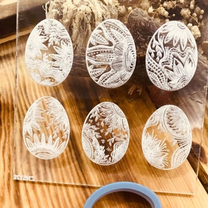 May include: A set of seven clear acrylic Easter egg stamps with intricate white floral designs. The stamps are arranged in a circle on a wooden surface. A blue silicone oval ring is also visible in the image.