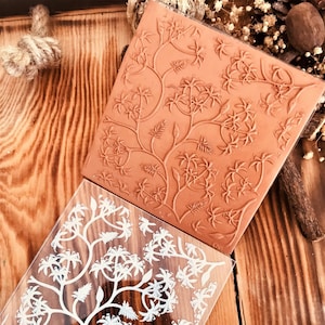 Puede incluir: Dos sellos de acrílico transparente con un diseño floral. El sello superior es marrón y el sello inferior es blanco.