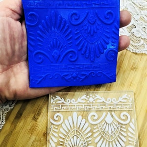 May include: A blue silicone stamp and a clear acrylic stamp with a repeating Greek key and floral design. The blue stamp is held in a hand. The clear stamp has the text "CNS044".