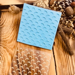 May include: A blue silicone stamp with a repeating pattern of dolphins. The stamp is on a clear plastic sheet with the text "CNS 333" printed on it.