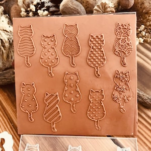 Peut inclure: Un ensemble de 10 tampons en acrylique transparent avec différents motifs de chats. Les tampons sont disposés en deux rangées sur une surface brune. Les tampons sont conçus pour être utilisés avec de l'encre et du papier pour créer des objets artisanaux personnalisés.