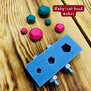 May include: A blue silicone mold with three hexagonal cavities for making ruby-cut beads. The mold is attached to a wooden surface with screws. There are several small, red and green beads scattered around the mold. The text "Ruby-cut bead maker" is written in red on a white background.