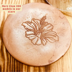 Puede incluir: Un posavasos redondo marrón con un diseño de flor de hibisco blanco. El texto "Más de 300 modelos en nuestra tienda" es visible en la esquina superior izquierda de la imagen.