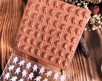 Könnte beinhalten: Ein brauner Silikonstempel in Form eines Lebkuchenmanns mit einer durchsichtigen Acrylplatte mit weißen Lebkuchenmann-Umrandungen. Der Stempel befindet sich auf einer Holzoberfläche mit natürlichen Elementen.