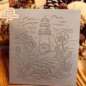 Puede incluir: Un sello de goma gris con un diseño de faro y flores. El sello mide 7-7cm, 10-10cm y 14-14cm. El texto "Rubber Texture Mat" está impreso en el sello.