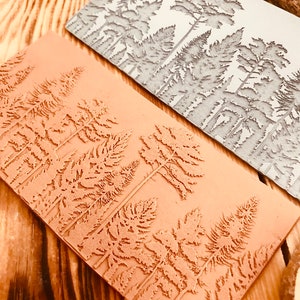 May include: Two rectangular rubber stamps with a forest design. The top stamp is grey and the bottom stamp is brown. Both stamps feature a repeating pattern of stylised pine trees.
