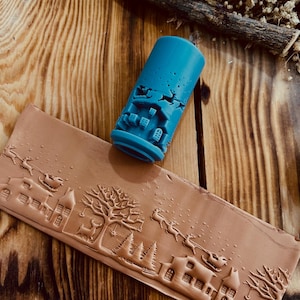 Op de afbeelding: Een blauwe rol met een kerstscèneontwerp. De rol wordt gebruikt om een reliëfpatroon op klei of fondant te maken. Het ontwerp toont een huis, een boom, rendieren en een slee.