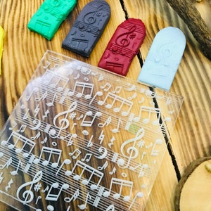 K&ouml;nnte beinhalten: Eine Sammlung farbiger, rechteckiger Stempel mit Musikmotiven und eine transparente Acrylplatte mit eingravierten Noten. Die Stempel sind gr&uuml;n, grau, rot und hellblau. Der Hintergrund ist eine Holzoberfl&auml;che.