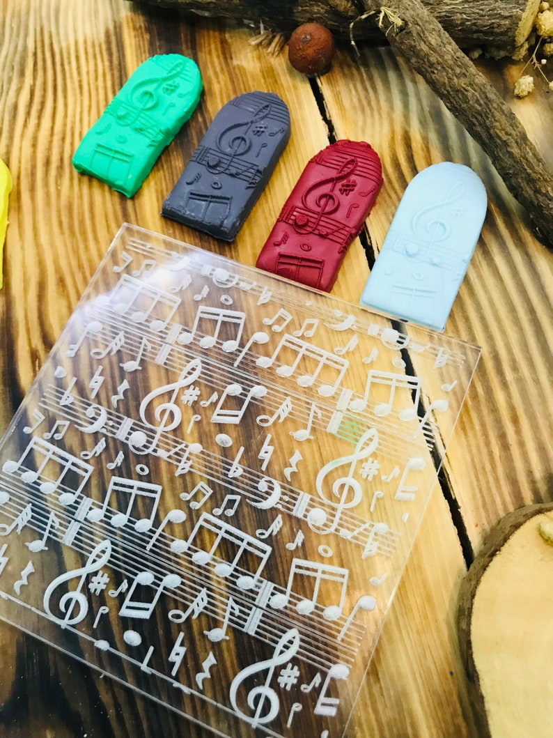 K&ouml;nnte beinhalten: Eine Sammlung farbiger, rechteckiger Stempel mit Musikmotiven und eine transparente Acrylplatte mit eingravierten Noten. Die Stempel sind gr&uuml;n, grau, rot und hellblau. Der Hintergrund ist eine Holzoberfl&auml;che.