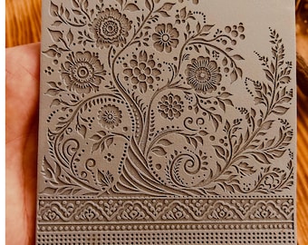 Könnte beinhalten: Eine 10 x 10 cm große Texturmatte aus Gummi mit einem detaillierten Blumenmuster. Die Matte zeigt ein sich wiederholendes Muster aus Blättern, Blumen und Spiralen. Das Muster ist in das Gummi geprägt und erzeugt eine erhabene Textur.