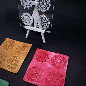 May include: A clear plastic sheet with four different mandala designs. The sheet is on a white easel. Below the sheet are four squares of clay with the mandala designs embossed in them. The clay squares are in different colors: gold, red, green, and orange.