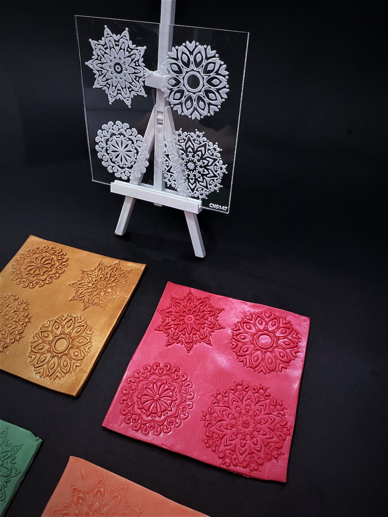 May include: A clear plastic sheet with four different mandala designs. The sheet is on a white easel. Below the sheet are four squares of clay with the mandala designs embossed in them. The clay squares are in different colors: gold, red, green, and orange.