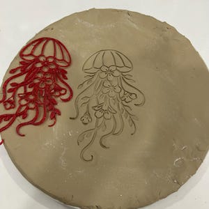 May include: A round, tan clay piece with a jellyfish design. A red, detailed jellyfish stamp is on the left. The stamped jellyfish design is in the center. The jellyfish has a bell-shaped top and flowing tentacles with floral details.