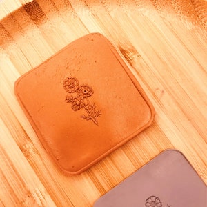 May include: Two square coasters with floral designs. One is a terracotta color, and the other is a muted purple. Both coasters have a pressed floral design. The coasters are on a bamboo tray.