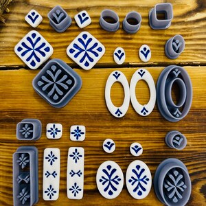 May include: A set of 20 polymer clay cutters in various shapes and sizes, including squares, ovals, and rectangles. The cutters are gray and feature a snowflake design. The set also includes several white polymer clay pieces with blue snowflake designs.