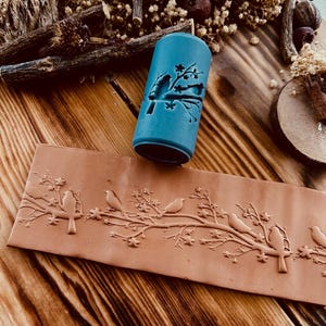 May include: A blue rolling pin with a design of birds on a branch. The rolling pin is used to create an embossed design on clay or dough. The design is also shown on a piece of clay.