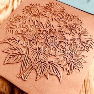 May include: A close-up of a terracotta-colored clay stamp with a design of five sunflowers with detailed leaves. The stamp is on a wooden surface.