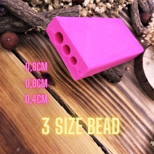 Puede incluir: Herramienta de fabricación de cuentas de plástico rosa con tres agujeros de diferentes tamaños: 0,8 cm, 0,6 cm y 0,4 cm. El texto "3 SIZE BEAD" está impreso en una superficie de madera.