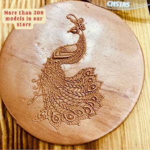 Puede incluir: Un azulejo de arcilla redondo de color cobre con un diseño de pavo real detallado en relieve. El azulejo tiene un diámetro de aproximadamente 15 cm. El texto "Más de 300 modelos en nuestra tienda" está impreso en una etiqueta amarilla en la esquina superior izquierda de la imagen.