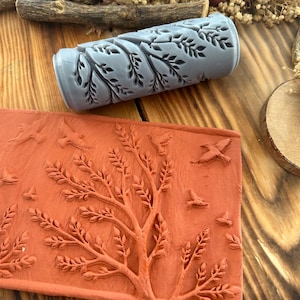 Può includere: Un rullo grigio con un motivo a foglie e un timbro rettangolare color terracotta con un disegno di albero e uccelli. Gli strumenti sono probabilmente per l'artigianato o scopi decorativi, con motivi ispirati alla natura.