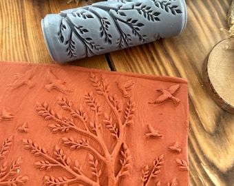 May include: A gray, cylindrical roller with a leaf pattern and a rectangular, terracotta-colored stamp with a tree and birds design. The tools are likely for crafting or decorative purposes, featuring nature-inspired motifs.