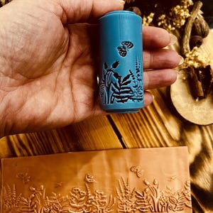 Könnte beinhalten: Ein blauer Gummistempel mit einem Blumen- und Schmetterlingsdesign. Der Stempel wird in einer Hand gehalten und das Design wurde auf ein Stück Leder gestempelt.