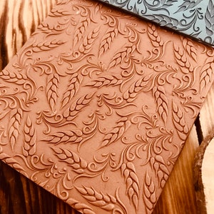 May include: Two textured rolling pins with a wheat and floral design. The rolling pins are brown and grey and are on a wooden surface.