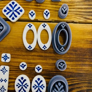May include: A set of 18 cookie cutters in various shapes and sizes, including circles, ovals, squares, and a snowflake design. The cutters are made of a light gray plastic and are arranged on a wooden surface.