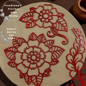 May include: A close-up of a clay stamp set with red floral designs. The set includes two large flower stamps, a decorative stem, and the text "FlexStamp Flexible Clay Stamp" and "KINDLY WATCH THE VIDEO". The stamps are on a light-colored surface.
