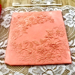 May include: A pink silicone stamp with a floral design. The design features a border of flowers and leaves around the edge of the stamp.