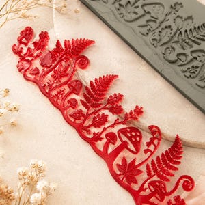 May include: A translucent red silicone mould featuring a detailed botanical design of ferns, various mushrooms, and small leaves, accompanied by a matching grey rectangular silicone mould with the same intricate forest-themed relief pattern.