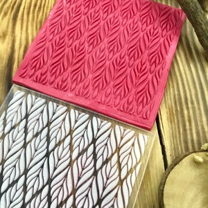 May include: A close-up of a red silicone stamp with a repeating pattern of white leaves. The stamp is on top of a clear acrylic sheet with the same pattern.