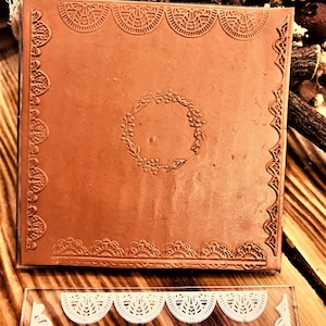 Op de afbeelding: Een bruine kleitegel met een decoratieve rand en een ontwerp van een bloemenkrans in het midden. De tegel ligt op een houten oppervlak.