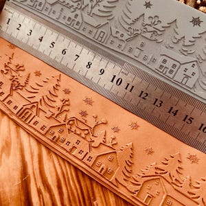 May include: A close-up of a grey rubber stamp featuring a winter village scene. The stamp is positioned on a wooden surface alongside a ruler marked in millimetres and inches. The stamp depicts a row of houses with snow-covered roofs, trees, and snowflakes.