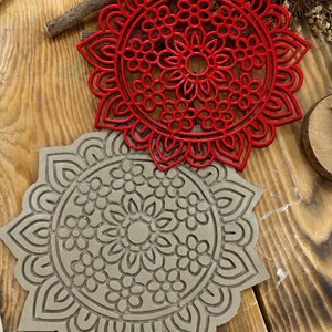 May include: Two circular coasters featuring an intricate floral mandala pattern. One coaster is a vibrant red plastic with a raised openwork design, while the other is a solid light grey material with the same floral mandala pattern embossed into it.